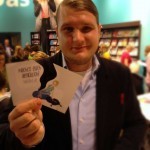 Viermalbe am Stand des frechverlags auf der Buchmesse: hält eine zerschnitte Postkarte hoch